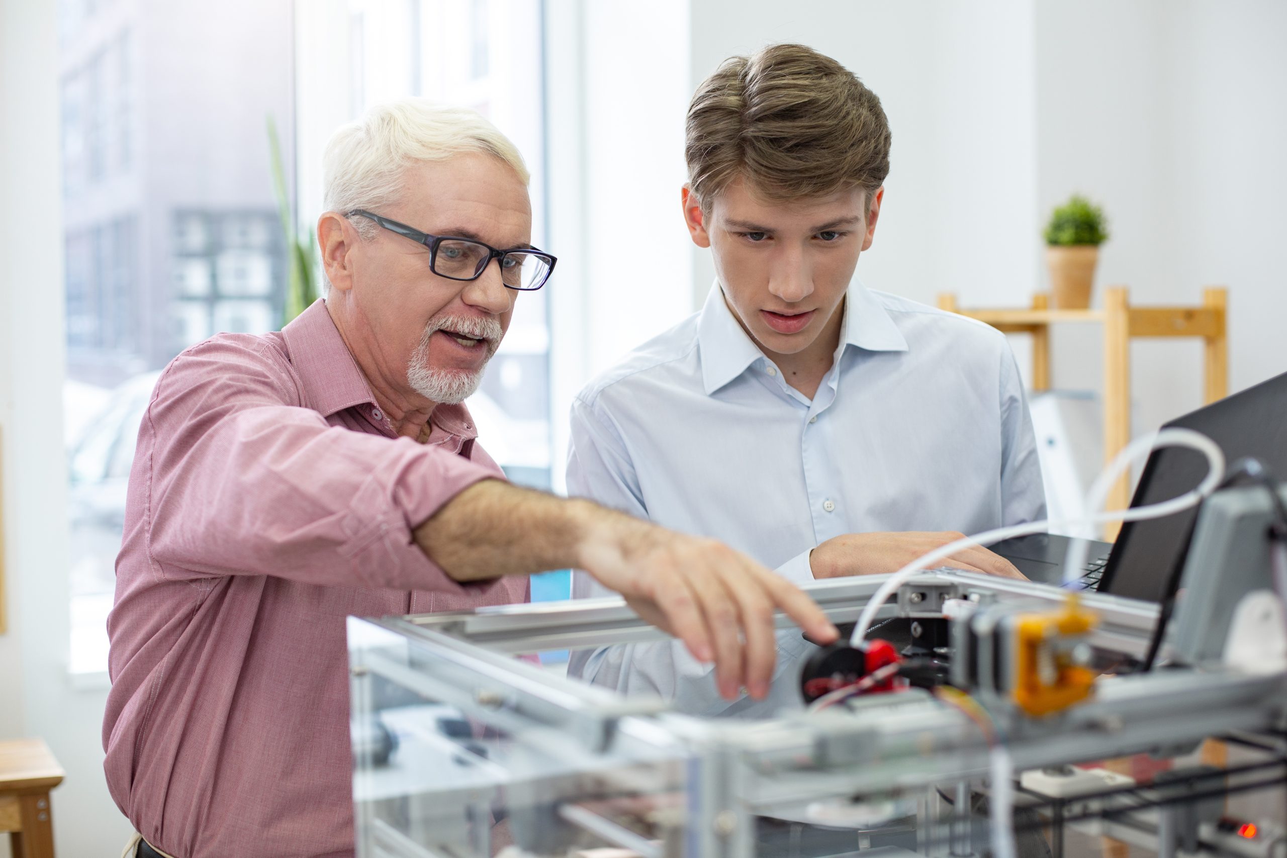 Sharing knowledge. Experienced senior engineer instructing his young intern about 3D printers while pointing at the important parts of mechanism